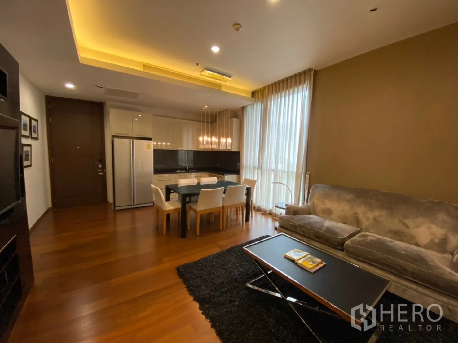 公寓 出租在Watthana, Bangkok - 客厅配灰色沙发、黑色地毯、木地板与大窗，旁接厨房。