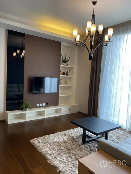 Condominium for rent in Watthana, Bangkok - Living room with wall‑mounted TV, built‑in shelving, chandelier, shag rug and large window.