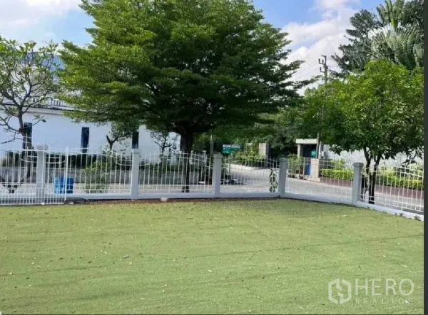 Detached House for rent in Samut Prakan - Corner lawn with white metal fence facing a quiet tree‑lined street.