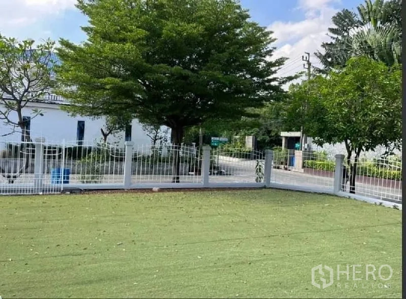 Detached House for rent in Samut Prakan - Corner lawn with white metal fence facing a quiet tree‑lined street.