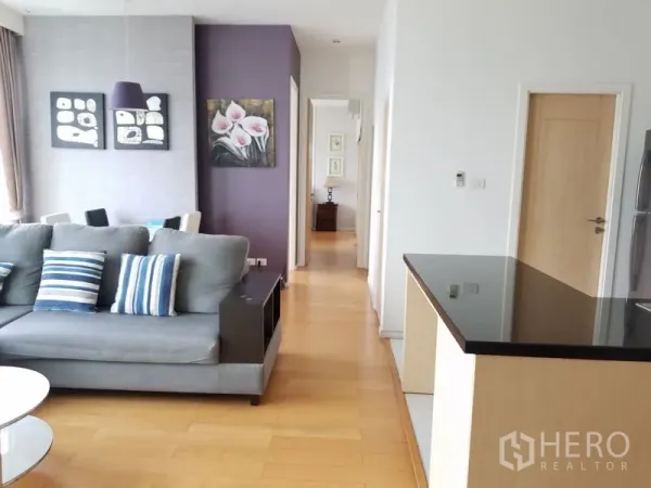 Condominium for rent in Chatuchak, Bangkok - Bright open‑plan living and dining area with a grey sofa, purple accent wall and a hallway beside the kitchen island.