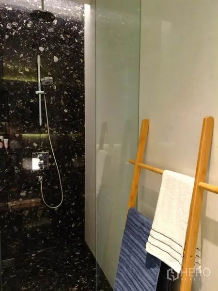 Condominium for rent in Khlong Toei, Bangkok - Glass-enclosed shower with rainfall head and dark stone wall, towels on wooden ladder.