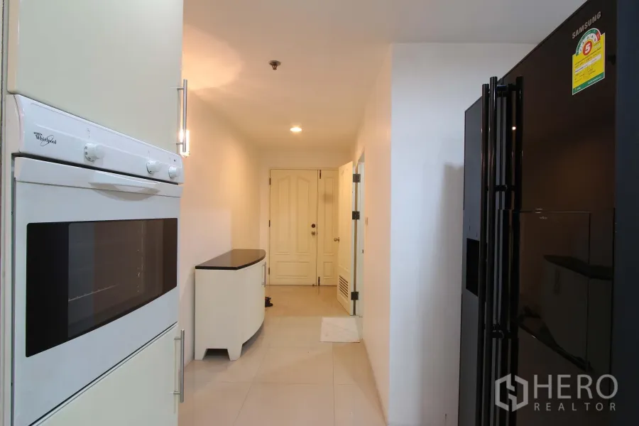 公寓 出租在Watthana, Bangkok - 明亮的走廊式厨房，带嵌入式烤箱、黑色对开门冰箱及通往入户门的过道。