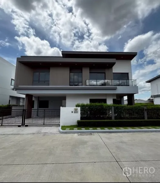 独立式住宅 出租或出售在Lat Krabang, Bangkok - 现代两层独栋别墅，玻璃阳台、入户庭院与整齐绿篱，天空多云。