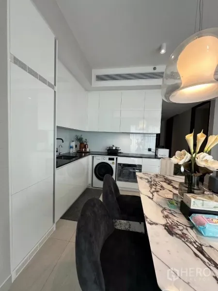 Condominium for rent in Bang Rak, Bangkok - Sleek U-shaped white kitchen with built-in oven, cooktop, and washer beside a marble breakfast bar and pendant light.