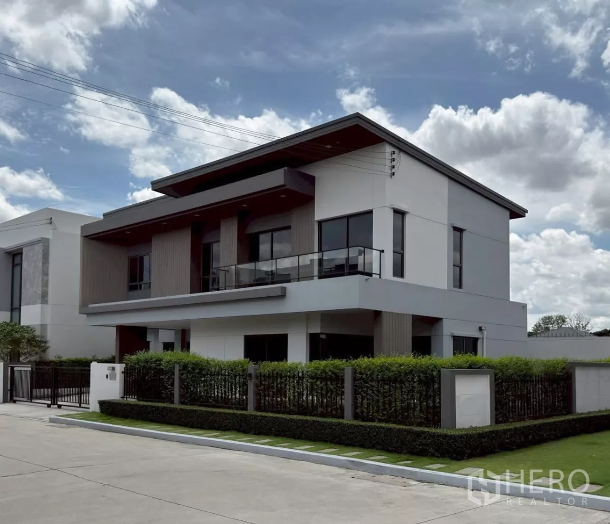 独立式住宅 出租或出售在Lat Krabang, Bangkok - 转角视角的独栋住宅，带玻璃栏杆阳台、大窗户与景观绿篱围栏。