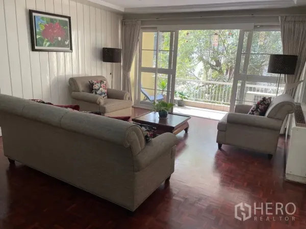 Condominium for rent in Watthana, Bangkok - Sunlit living room with sofa set, coffee table and sliding doors opening to a leafy balcony.