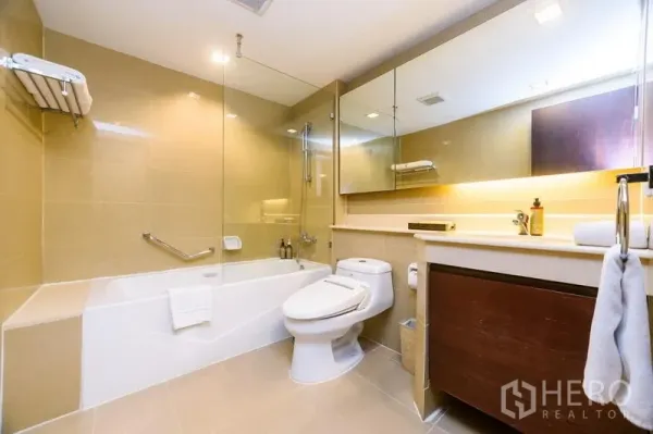 Condominium for rent in Khlong Toei, Bangkok - Modern bathroom with bathtub, glass shower screen, large mirror and long vanity countertop.