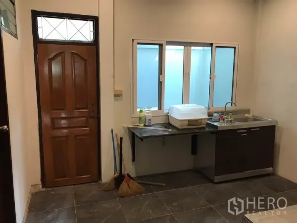 Townhouse for rent or sale in Phra Khanong, Bangkok - Secondary kitchen area with sink, sliding window, wooden door and tiled floor.