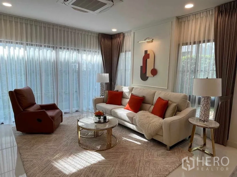 Detached House for rent in Saphan Sung, Bangkok - Sunlit living room with cream sofa, orange cushions, recliner and round coffee table by floor-to-ceiling windows.