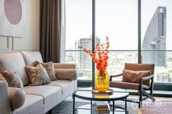 Condominium for rent in Khlong Toei, Bangkok - Contemporary living room with sofa and armchair beside large windows framing a city skyline view.