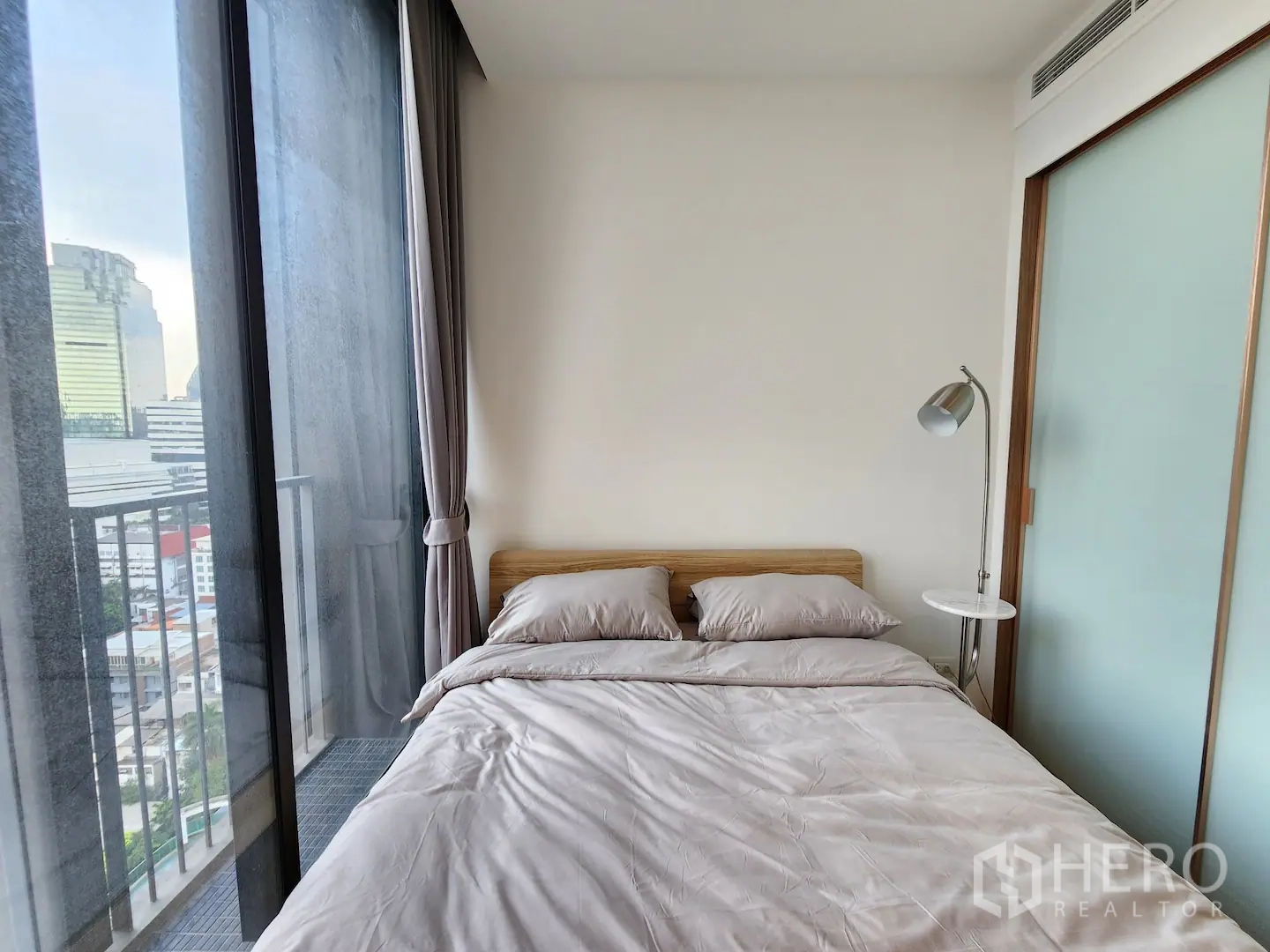 Condominium for rent in Watthana, Bangkok - Compact second bedroom with bed by a tall balcony window and reading lamp, city skyline beyond.