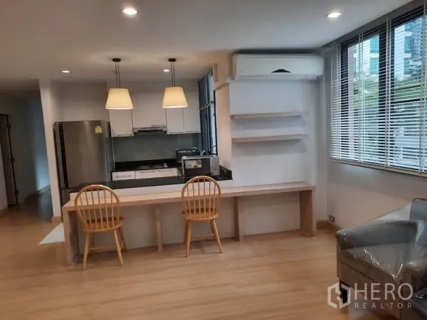 Condominium for rent in Watthana, Bangkok - Open kitchen with black countertop, stove and stainless refrigerator, plus a long built-in desk under windows and wall shelves.