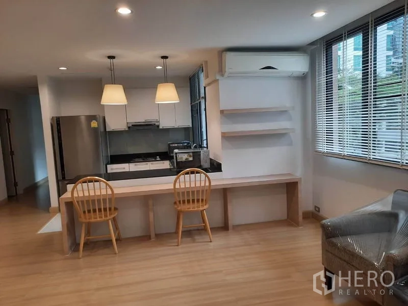 Condominium for rent in Watthana, Bangkok - Open kitchen with black countertop, stove and stainless refrigerator, plus a long built-in desk under windows and wall shelves.