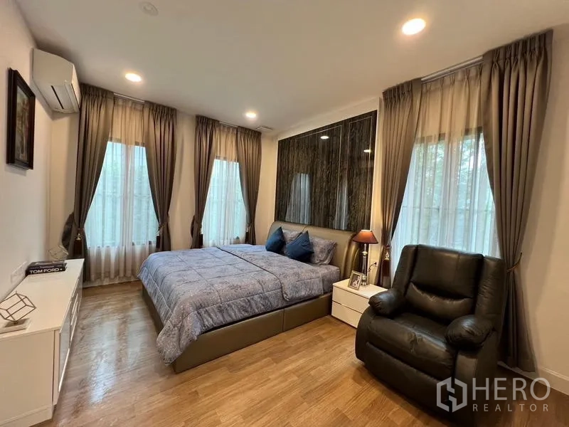 Detached House for rent or sale in Saphan Sung, Bangkok - Spacious bedroom with tall windows, a recliner chair and bedside tables.