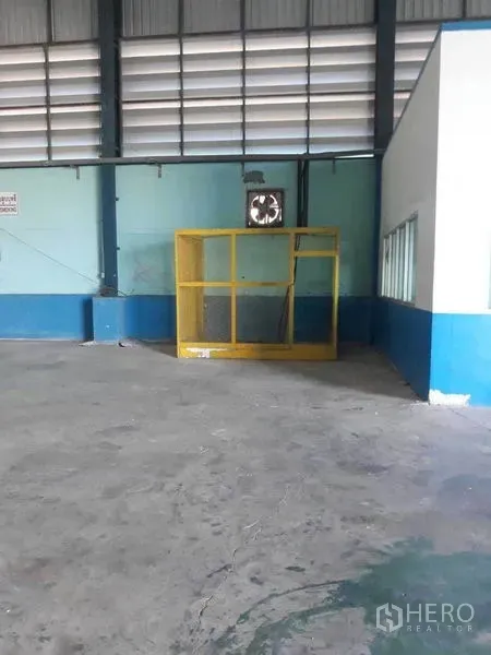 Factory for rent in Samut Prakan - Warehouse bay featuring a yellow safety cage, wall ventilation fan, and adjacent office windows.
