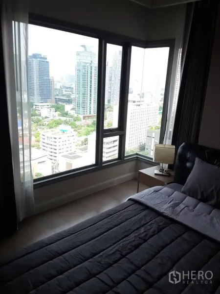 Condominium for rent in Khlong Toei, Bangkok - Corner bedroom with floor-to-ceiling windows offering a Bangkok skyline view and a bedside table with lamp.