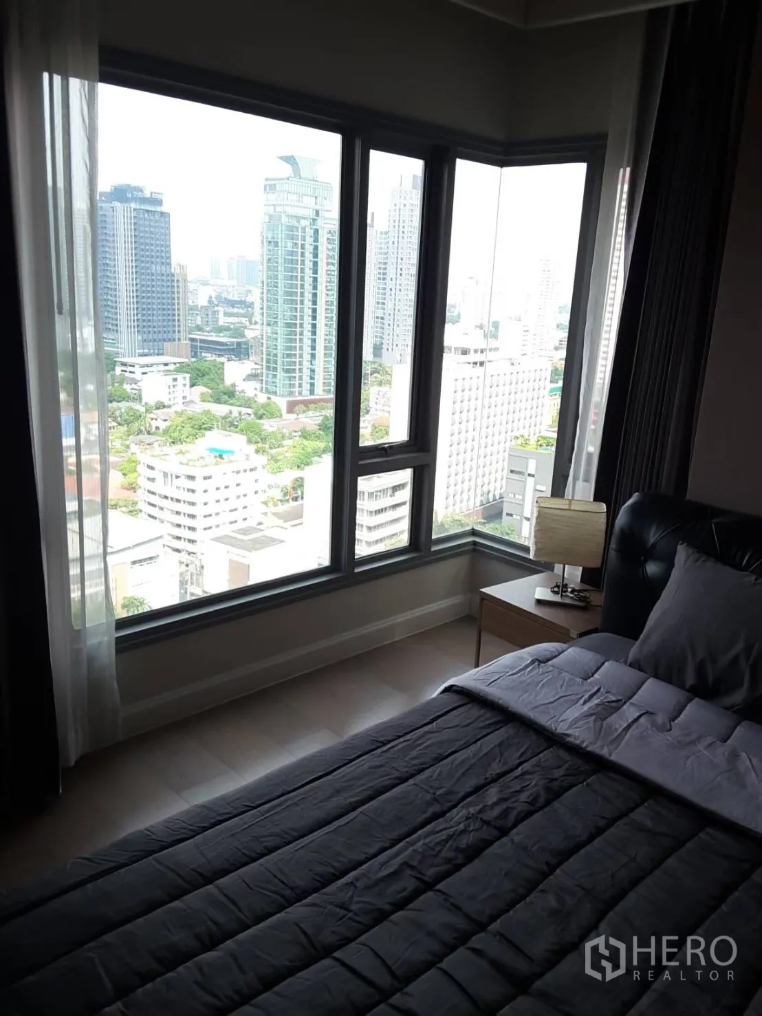 Condominium for rent in Khlong Toei, Bangkok - Corner bedroom with floor-to-ceiling windows offering a Bangkok skyline view and a bedside table with lamp.