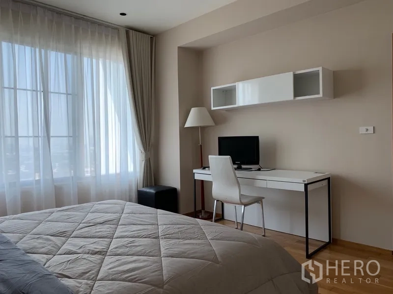 Condominium for rent in Khlong Toei, Bangkok - Bedroom with large window, sheer curtains and study desk with wall shelves.