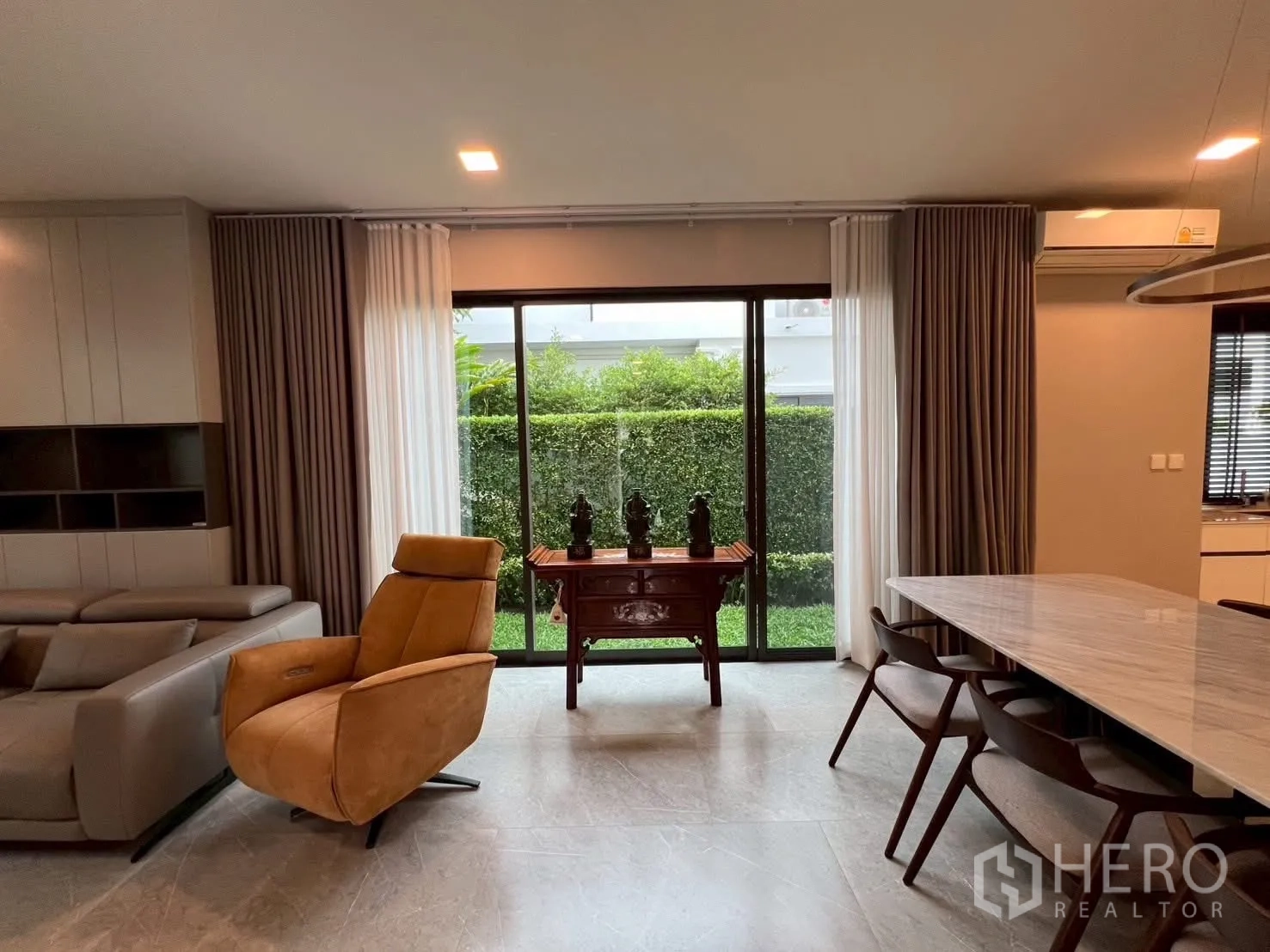 Detached House for rent in Samut Prakan - Bright lounge with sliding doors, curtains, and a tan recliner facing greenery.