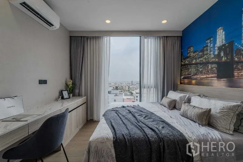 Condominium for rent in Pathum Wan, Bangkok - Bedroom with built-in desk and large window overlooking the Bangkok skyline, soft gray bedding.