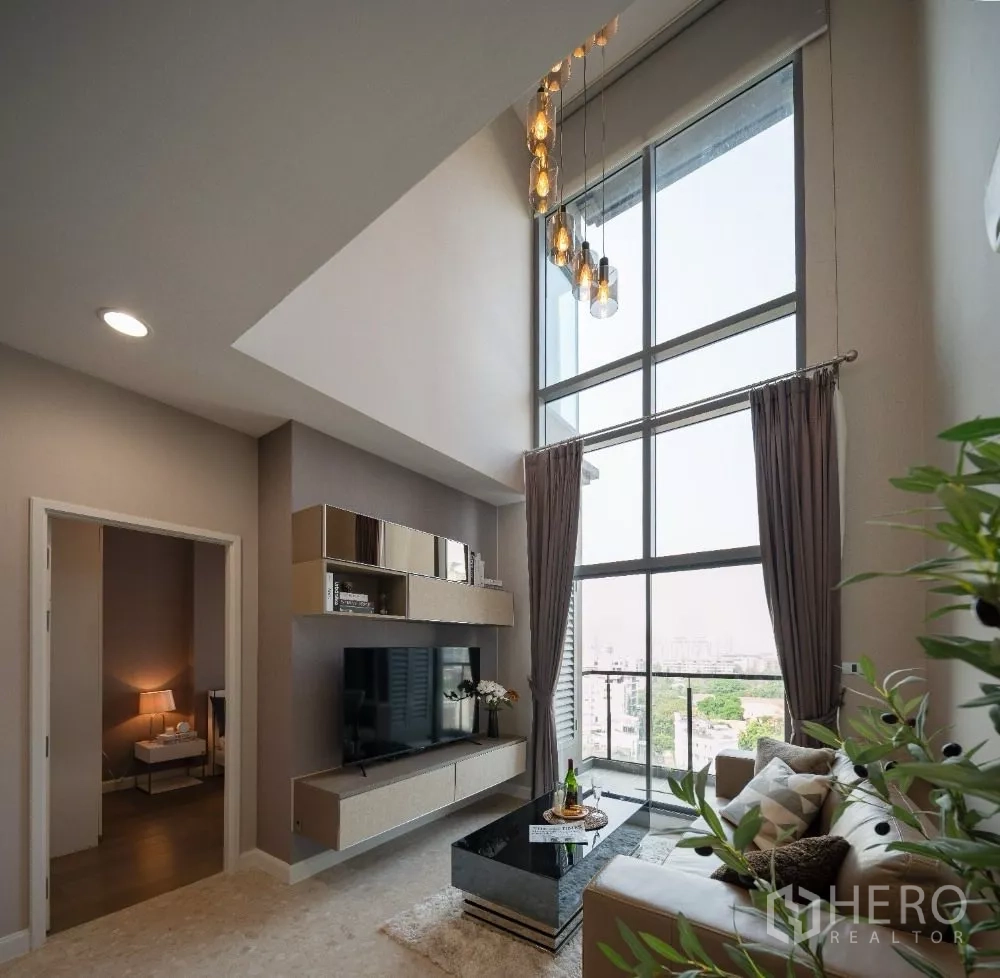 Condominium for rent or sale in Khlong Toei, Bangkok - High-ceiling living room with TV wall, large windows and a city-facing balcony.