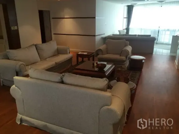 Condominium for rent in Khlong Toei, Bangkok - Open-plan lounge with multiple beige sofas and armchairs, large windows and wooden floors.