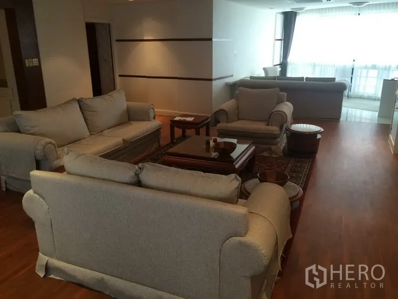 Condominium for rent in Khlong Toei, Bangkok - Open-plan lounge with multiple beige sofas and armchairs, large windows and wooden floors.