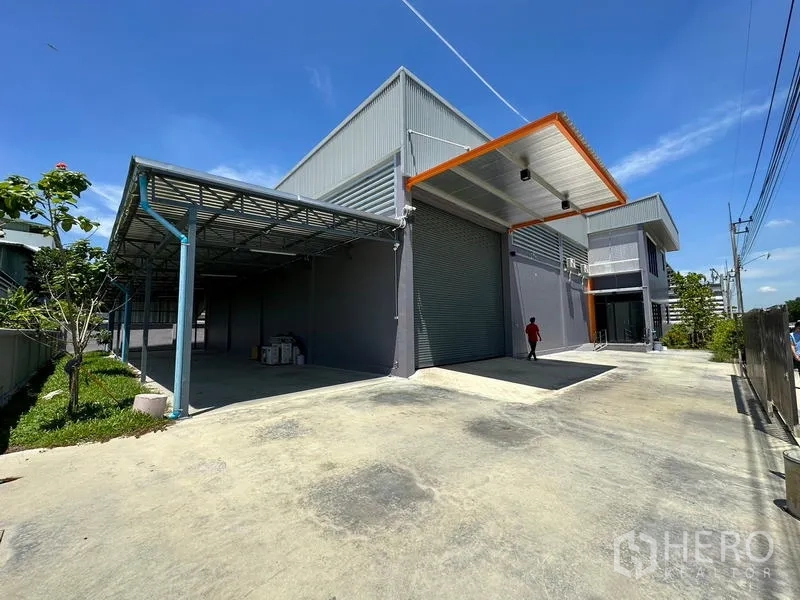 工厂 出租或出售在Lam Luk Ka, Pathum Thani - 工厂外观另一视角，卷闸门、雨棚与宽阔场地一览。