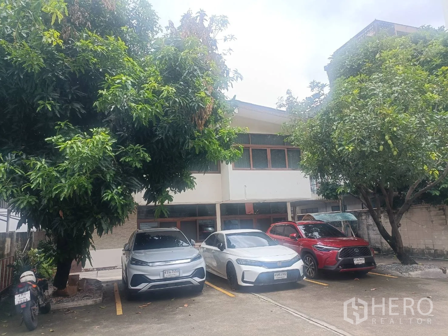 独立式住宅 出租在Bang Rak, Bangkok - 前院停车区，三辆车停在树荫下，对面是房子。