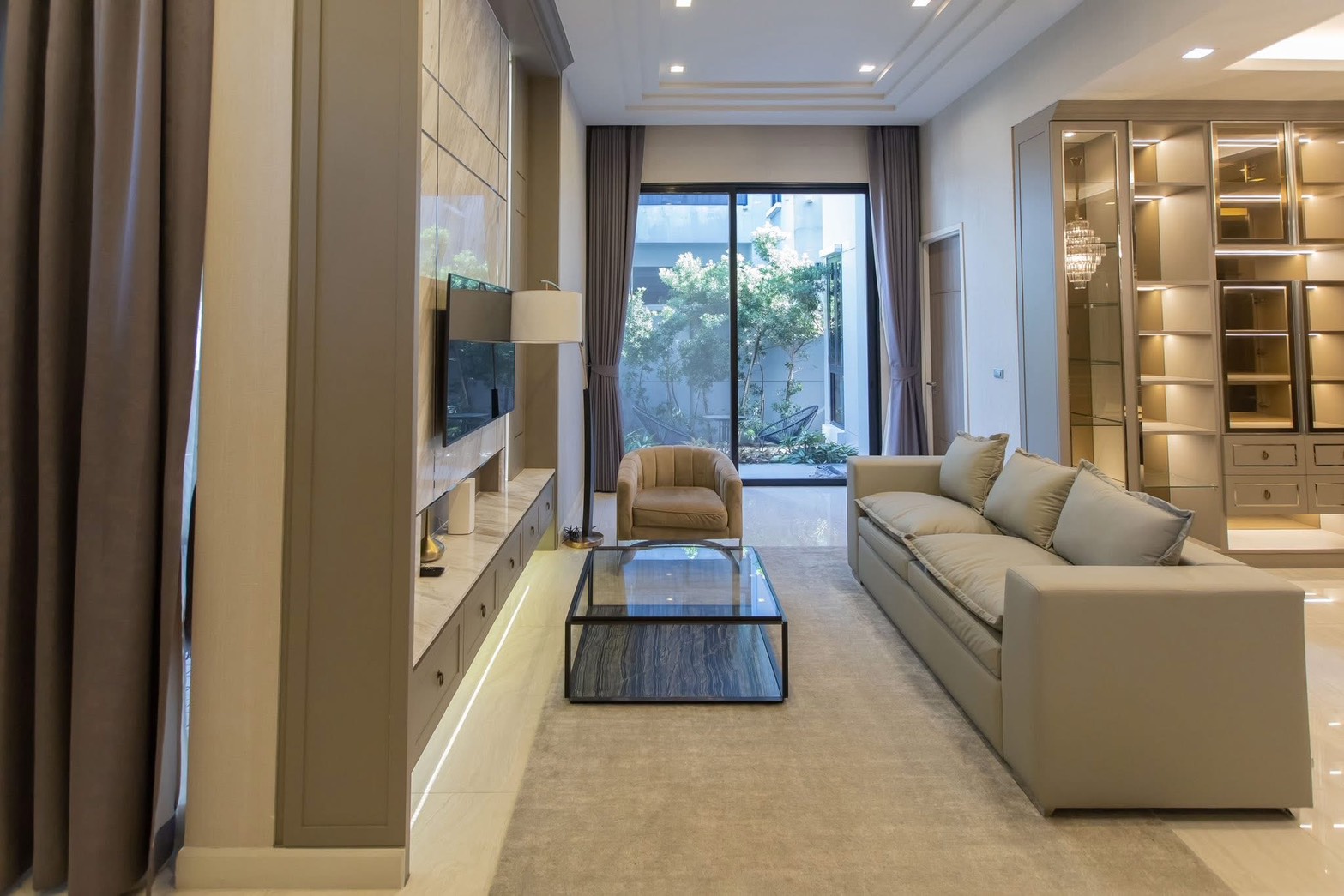 Detached House for rent in Lat Phrao, Bangkok - Bright living room with long sofa, TV wall panel, glass coffee table, and sliding doors to a small garden.