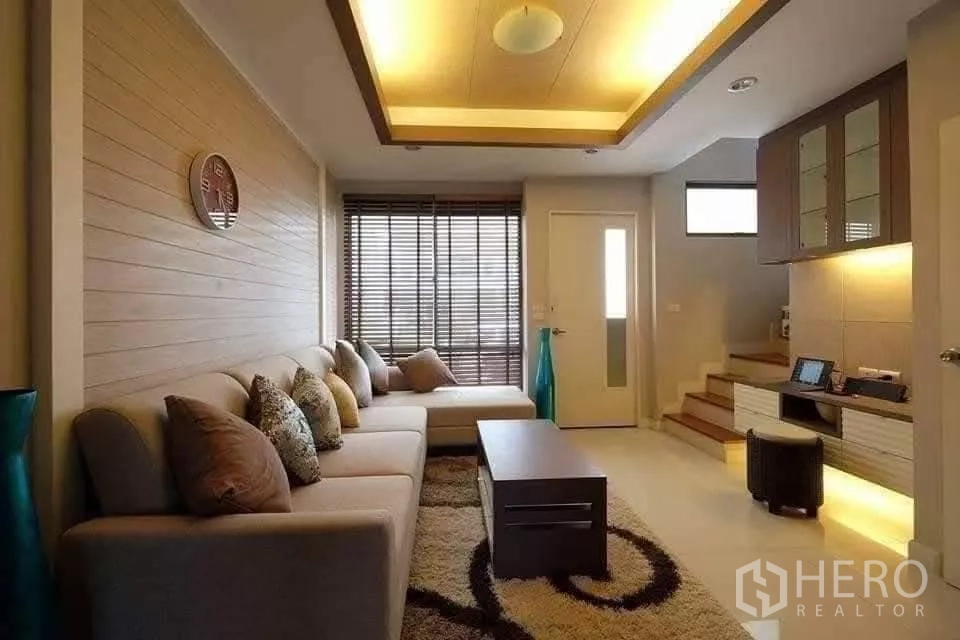 Townhouse for rent in Khlong Toei, Bangkok - Cozy lounge with L-shaped sofa, feature ceiling lighting, blinds, built-in cabinetry and stairs.
