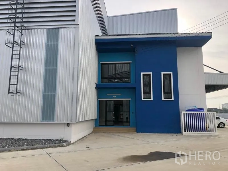 Factory for rent in Samut Prakan - Side entrance showing blue office façade attached to a modern metal‑clad factory building.
