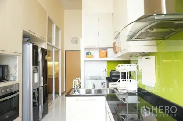 公寓 出租或出售在Watthana, Bangkok - 明亮的走廊式厨房，绿色玻璃防溅板、嵌入式烤箱、高柜与双开门冰箱。