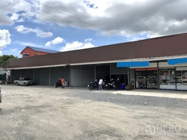 货栈 出租在Lam Luk Ka, Pathum Thani - 临街单层仓库排屋，配多扇卷帘门与宽阔碎石停车区，旁有小型店面。