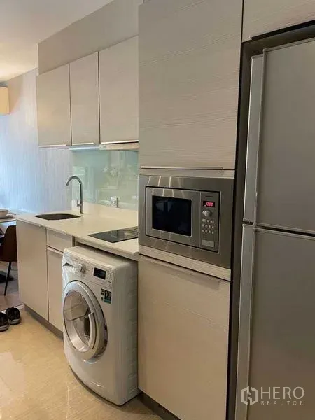 Condominium for rent in Watthana, Bangkok - Compact fitted kitchen with white cabinetry, sink, microwave, washing machine, and refrigerator.