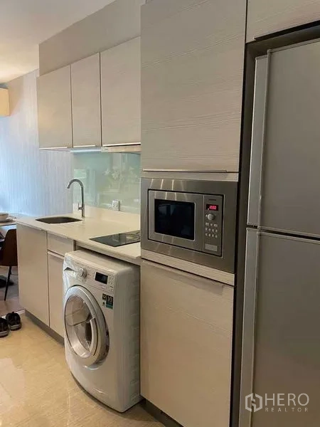 Condominium for rent in Watthana, Bangkok - Compact fitted kitchen with white cabinetry, sink, microwave, washing machine, and refrigerator.