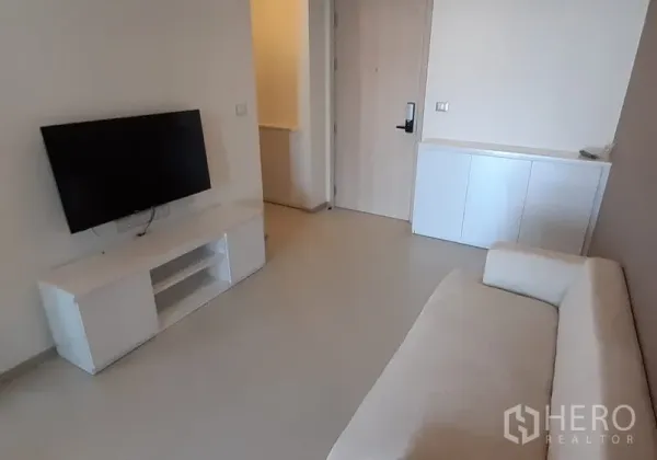 Condominium for rent in Khlong Toei, Bangkok - Minimalist living space with flat-screen TV on a white console and the entry door.