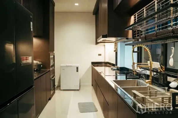 Detached House for sale in Sai Mai, Bangkok - Sleek galley kitchen with dark cabinetry, stone counters, double sink and a gold pull‑down faucet.