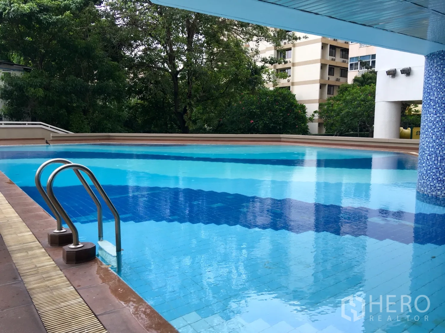 Condominium for rent or sale in Watthana, Bangkok - Outdoor swimming pool with stainless ladder, shaded deck and leafy surroundings.