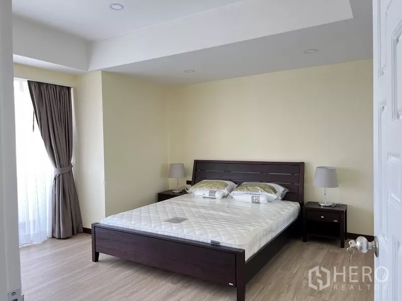 Condominium for rent in Khlong Toei, Bangkok - Master bedroom with dark wood bed, side tables with lamps and full-length curtains.