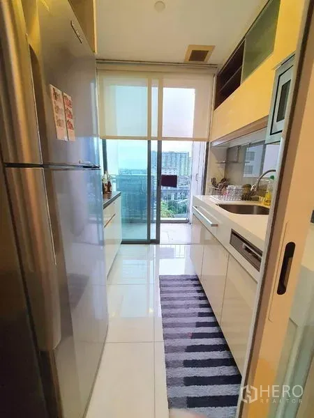 คอนโด ขายในBangkok - ครัวสไตล์แกลลีย์สีขาว ตู้บิลท์อิน ตู้เย็นสแตนเลส และประตูกระจกบานเลื่อนไปยังระเบียงวิวเมือง