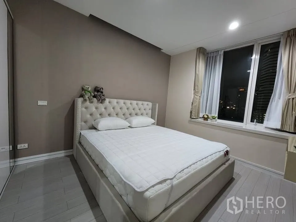 Condominium for rent in Huai Khwang, Bangkok - Master bedroom at night with upholstered bed and corner windows.