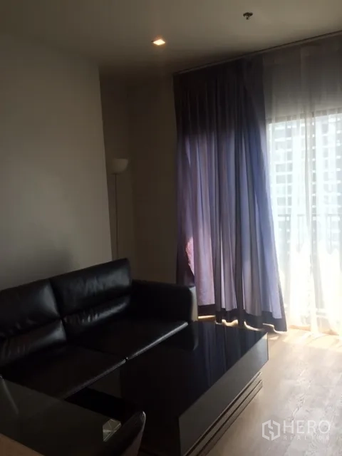 Condominium for rent in Bangkok - Cozy living room with L-shaped leather sofa, coffee table and tall curtains by sliding glass doors.