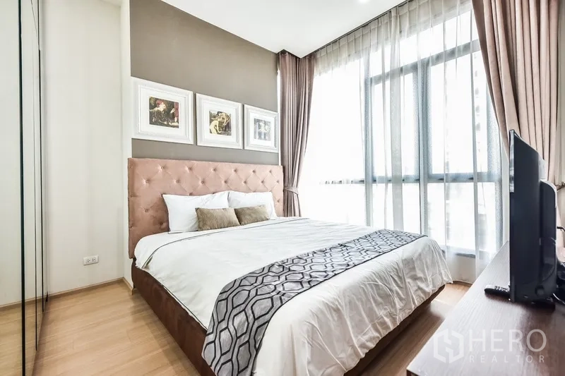 Condominium for rent or sale in Huai Khwang, Bangkok - Sunlit bedroom with tufted headboard, large window, TV, and built-in wardrobe.
