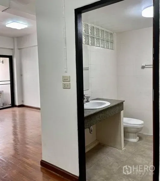 Home Office for rent in Khlong Toei, Bangkok - Bathroom entrance off a wood-floored room showing the sink, toilet and glass-block window.