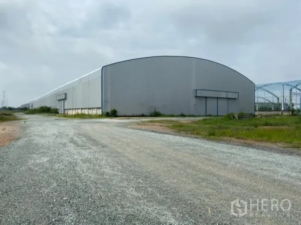 Warehouse for rent in Ban Pho, Chachoengsao - Large metal-clad warehouse exterior with wide gravel access road and oversized sliding loading doors.