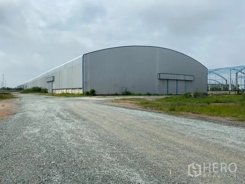 Warehouse for rent in Ban Pho, Chachoengsao - Large metal-clad warehouse exterior with wide gravel access road and oversized sliding loading doors.
