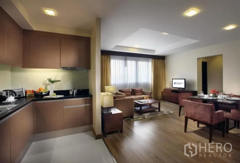 Condominium for rent in Khlong Toei, Bangkok - Modern L-shaped kitchen with wood cabinets and black countertop opening to a cozy living room and dining area with warm wood flooring.