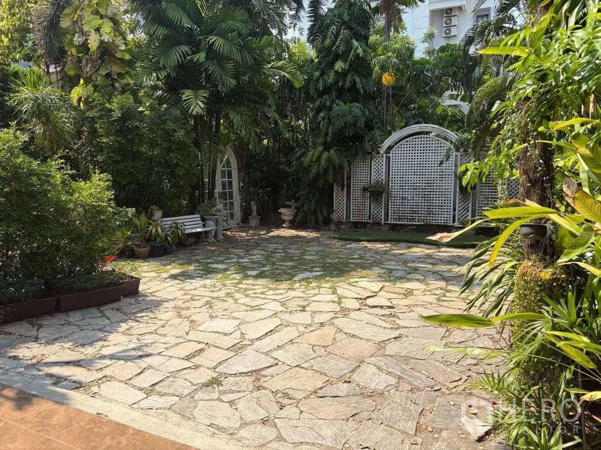 Detached House for rent in Phra Khanong, Bangkok - Stone-paved courtyard with a bench and lattice gate amid tropical plants.